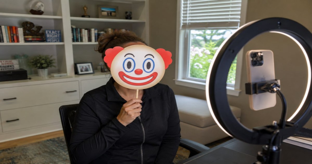 A person holds a clown emoji mask in front of their face while sitting at a desk with a ring light and phone